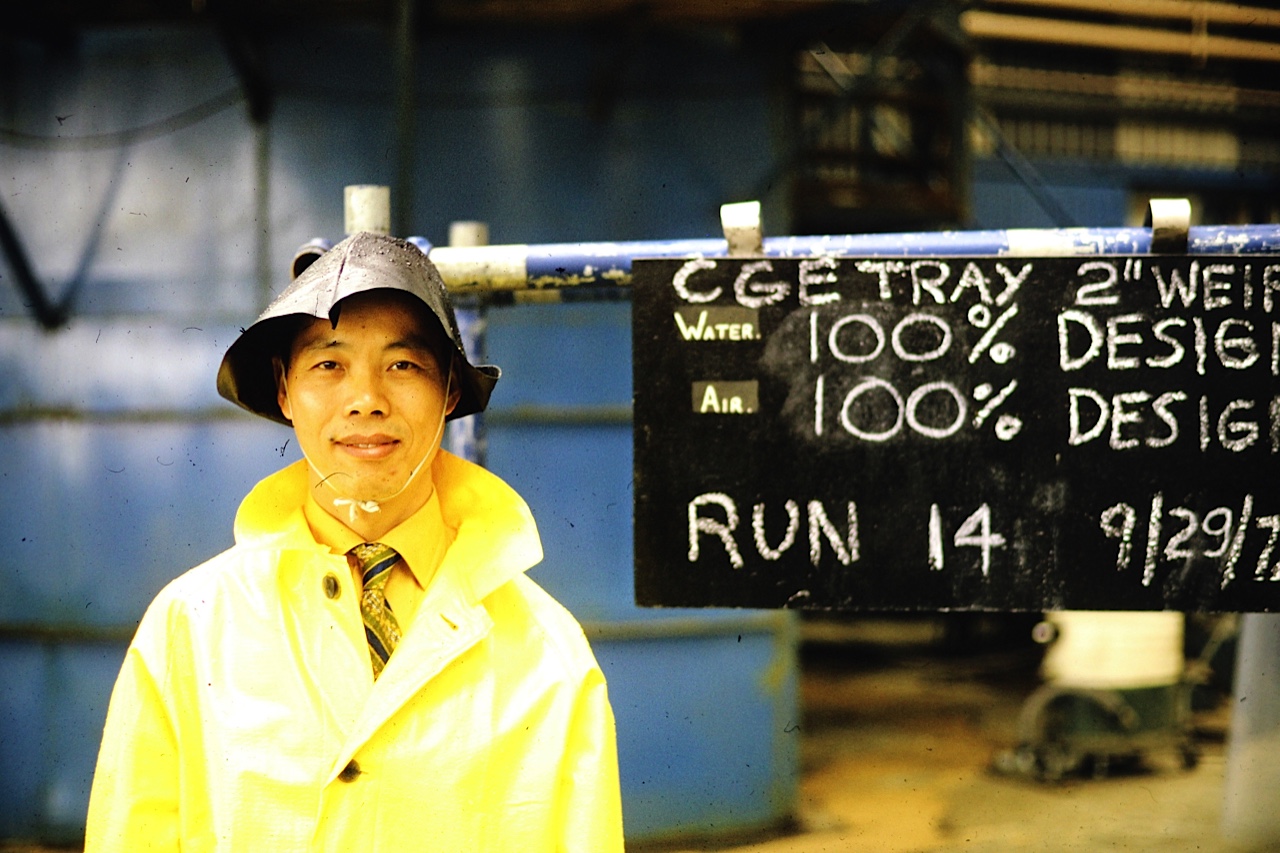 Shinn-Der Chang in a yellow rain coat at the GE heavy-water plant in Port Hawkesbury, Nova Scotia, in front of a chalkboard reading 'CGE TRAY 2" WEIR / WATER 100% DESIGN / AIR 100% DESIGN / RUN 14 9/29/73'.