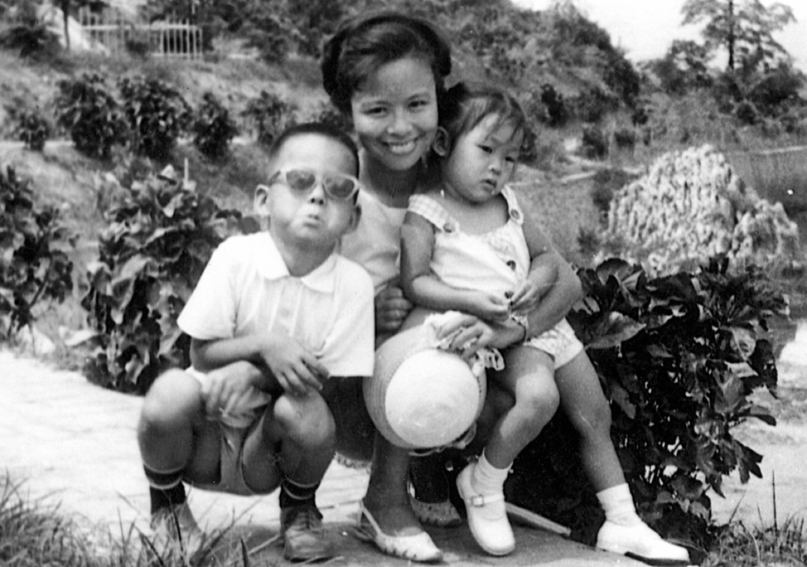 Rae with Nathan and Nancy as small children, sitting on a ledge in front of a garden, 1964
