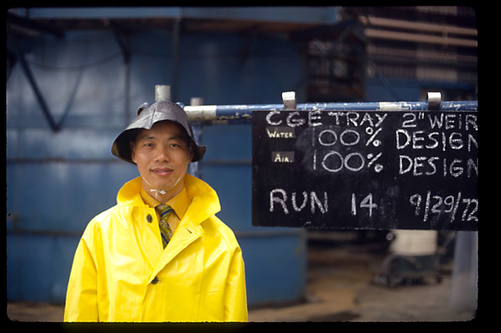 Shinn-Der Chang in a yellow rain coat and hat next to a chalkboard, 1972