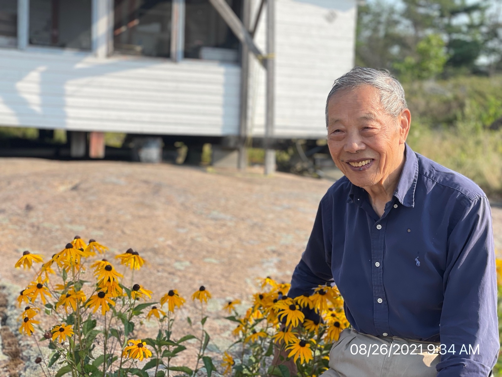 Shinn-Der Chang seated on the ground next to a flower bed, August 2021