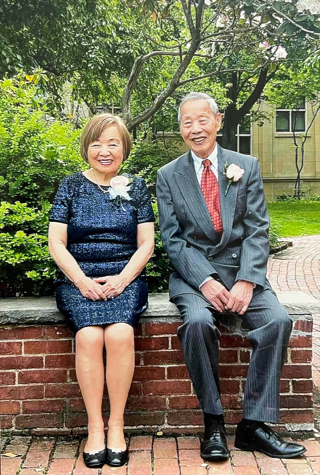 Rae and Shinn-Der seated together on a brick wall in front of a garden at Daniel and Joy's wedding, May 2022