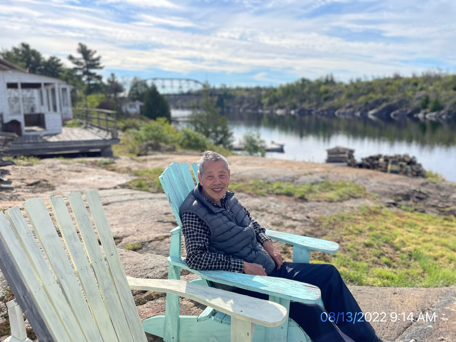 Shinn-Der Chang seated on a bench by the water at the cottage, August 2022